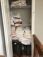 Closet with folded bed linens on shelves and black collapsible travel luggage at the bottom.