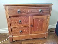 Front view of antique wood washstand showing top drawer, two lower drawers, and cabinet door with carved leaf pattern and metal ring pulls.