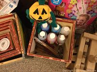 Photo of paint bottles including white, blue, and orange acrylic paints, paintbrushes, and a plush pumpkin toy inside a wooden box.