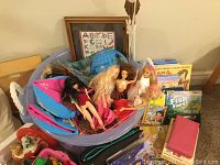 Photo of multiple Barbie dolls seated in large pink and blue plastic tub with big LEGO blocks, surrounded by various children's books stacked and laid out nearby.