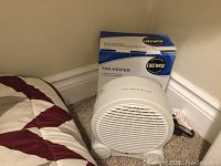 Small white Likewise brand fan heater in front of its box beside linens on carpet floor.