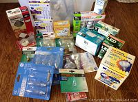 Assorted collection of boxed and loose light bulbs including appliance bulbs, flood lights, ampoules, and LED bulbs displayed on wooden floor.