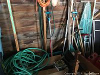 Photo showing coiled green garden hoses, multiple shovels and digging tools with blue handles, axe, and part of the ironing board in the background.