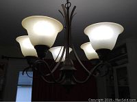 Lit chandelier showing five illuminated frosted glass shades in bell shape and bronze metal frame