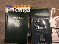 Religious books including a Holy Bible in green cover, another Holy Bible, a Catechism of the Catholic Church book, and pieces of the Adobe InDesign software manual.