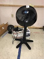 Photo showing large black Vornado pedestal fan and smaller Vornado table fan in background with basket of hair styling tools on floor.