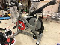 Full view of the silver frame spin bike with red and white star on the flywheel, showing padded handlebars and base with small wheels.