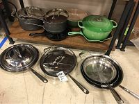Overview of cookware set showing Dutch oven, cast iron skillet, All Clad pans with lids, Cuisinart pot, and ceramic bowl on shelves and floor