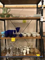 Full view of all glassware and ceramics on shelf including all cups, glasses, teacups, and bowl and pitcher.