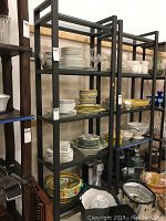 Two black metal frame shelving units with glass shelves, each loaded with various plates, bowls, and glassware for size reference.