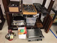 Overview of lot on wooden shelf showing chafing dish, flatware in wooden tray, plastic bin of utensils, grilling tools, Imperia box and Breville Smart Grill on floor.