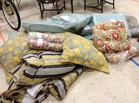 Assortment of outdoor seat cushions including floral prints, plastic wrapped cushions, striped cushions, and draperies.