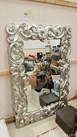 Front view of large rectangular mirror leaning against wall showing ornate frame with swirling patterns and reflective glass.