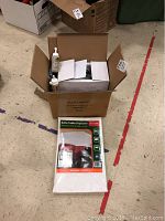 Box containing the airbrush kit components and instruction papers alongside the packaged sofa/table protector on floor.