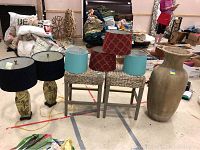 Photo showing two palm print lamps with shades (red and teal), two woven rattan stools, and a large handmade pottery vase on the right.