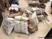 Wide view showing multiple quilts, pillows, cushions, folded curtains, and curtain rods grouped together on floor.