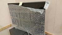 Front view of the Samsung TV wrapped in bubble wrap with stand attached, showing the screen and bezel.