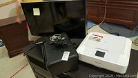 White Canon Pixma MG7720 printer with touchscreen, power cord, black HP Photosmart D110 all-in-one printer, and Samsung 40in TV behind them