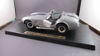 Side view of silver Shelby Cobra model car with driver-side door open, showing detailed interior and clear windshield. Mounted on black base with metal plaque label.