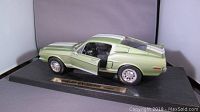 Side profile view of the green metallic 1968 Shelby GT-500KR die-cast model with white racing stripes, mounted on black display base with plaque. Driver door opened showing part interior.