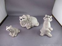 Three stone-look raccoon figurines arranged on gray background.