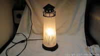 Front view of illuminated glass lighthouse lamp showing glass body, metal top and base, power cord with switch.