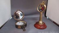 Brass Titanic engine room telegraph with wooden round base, dial with speed markings and two brass levers, standing beside vintage nautical compass with open lid showing mirror and compass face.