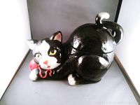 Full side angle view showing the black and white cat cookie jar with a red bow, curled and lying down with tail raised.