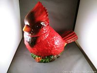 Front view of a bright red ceramic cardinal cookie jar with detailed feathers, yellow beak, and green and brown base depicting leaves and pine cones.
