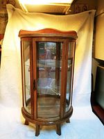 Small wooden curio cabinet with curved glass front panels on a white backdrop. Shows overall size and wood finish.