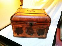 Closed wooden box showing three metal leaf decorations on front and one on lid, medium-brown polished wood.