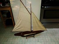 Full side view of a large wooden model sailboat with two fabric sails on a tiled floor.