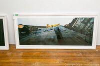 Full view of white-framed panoramic color photograph print depicting wooden pier planks and yellow crane against rocky background