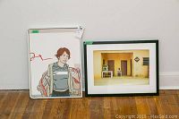 Framed illustrated portrait of a woman standing next to framed photographic print leaning against wall.