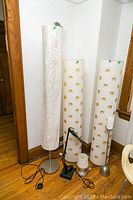 Tall white cylindrical floor lamp with crinkled fabric shade, next to two cream-colored cylindrical floor lamps with circular cutouts, a black adjustable desk lamp, a small bedside lamp with glass base, and a metal candlestick holder with candle