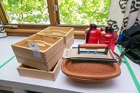 Overview of the lot showing 4 wooden holders, 2 red water bottles, the cast iron grill press, clay roaster, and plumbing snake on a table under window light.