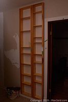 Two tall, narrow pine shelving units with open, adjustable shelves placed side by side against a wall next to a doorway.