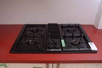 Full view of Jenn Air gas cooktop sitting on red countertop, showing four burners and central vent/grill with three knobs