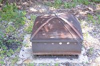 Top and angled views of square metal firepit with mesh cover and attached poker tool, sitting on paving stones.