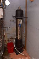 Full view of the Maytag water filtration unit installed vertically next to plumbing and wall with red bucket on floor.