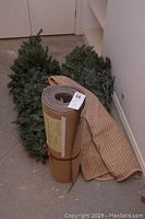 Artificial Christmas tree sections along with rolled rug pad and folded striped rug on the floor in a corner.