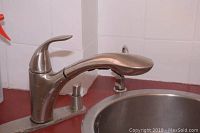 Close-up of stainless steel single-handle kitchen faucet with curved spout on red countertop with white tiled backsplash.