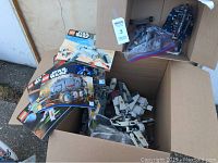 Box containing multiple LEGO Star Wars instruction manuals and various pieces, including a partially assembled starfighter model in white and gray.