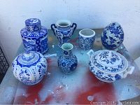 Full view of the seven Delft Blue pieces on surface showing diverse shapes including ginger jar, casserole, egg-shaped decor, and small vases.