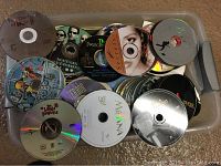 Top view of a plastic container filled with a variety of loose CDs and DVDs with visible titles including 'Moana', 'The Twilight Saga', and others, no cases included.