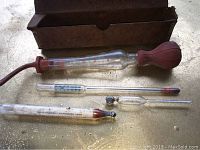 Photo showing four antique glass thermometers arranged beside a worn rectangular cardboard box on a flat surface. One thermometer has a red rubber bulb attached.