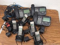 All phones and accessories arranged on a wooden table showing 3 desk top phones and 5 handheld phones with cords and power adapters.