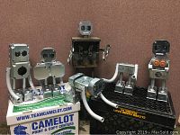 Photo showing all 6 handmade junk art robots arranged on boxes and crate against a brown wall.