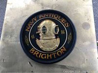 Circular navy antiques sign with raised brass diver's helmet and lettering against black background on a flat surface