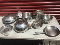 Overview of all stainless steel cookware items including pots, pans, colanders and racks laid out on a table.